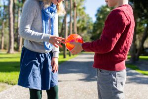 Two children standing outdoors on a sunny day, holding a bright orange and red ball together. The focus is on their hands and torsos; trees and a pathway are visible in the background. - Villas by the Sea Resort, Jekyll Island