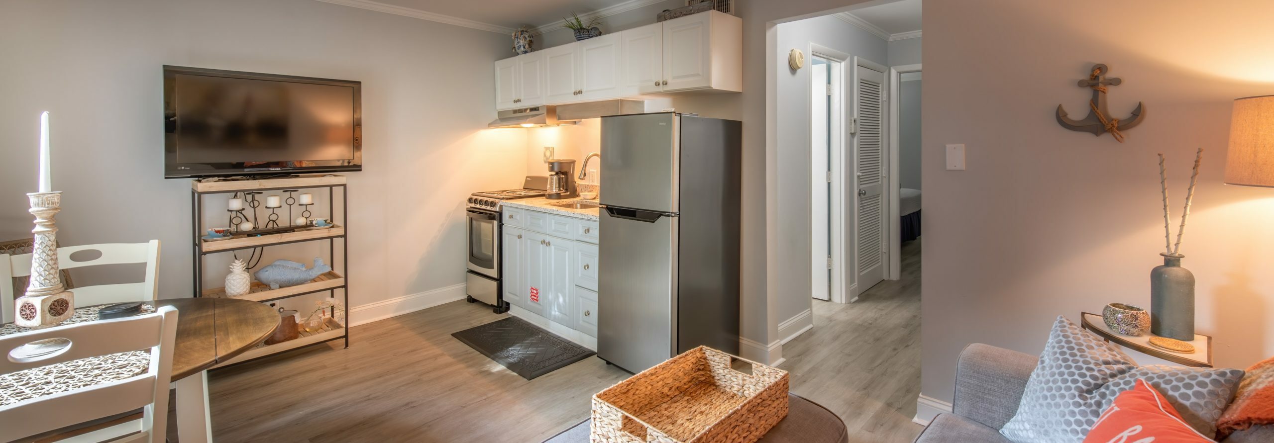 Small, cozy kitchen and living area with a TV, refrigerator, stove, dining table, and coastal decor. A hallway leads to other rooms. Warm lighting and neutral colors create a welcoming atmosphere. - Villas by the Sea Resort, Jekyll Island