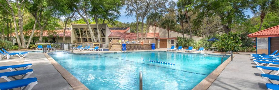 Outdoor swimming pool surrounded by blue lounge chairs and trees, with a modern clubhouse building in the background under a partly cloudy sky. - Villas by the Sea Resort, Jekyll Island